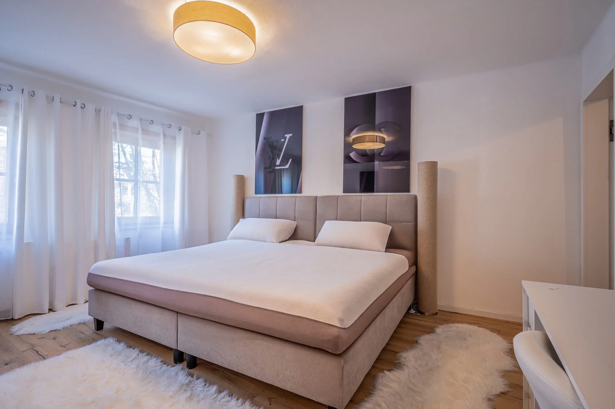 Helles Schlafzimmer mit gepolstertem Doppelbett, weißen Vorhängen, Fellen und Holzboden